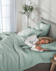 Woman lying in bed with light green bedding in a bright bedroom.