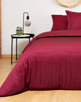 Burgundy bedding set on a bed in a room with wooden flooring and a side table.