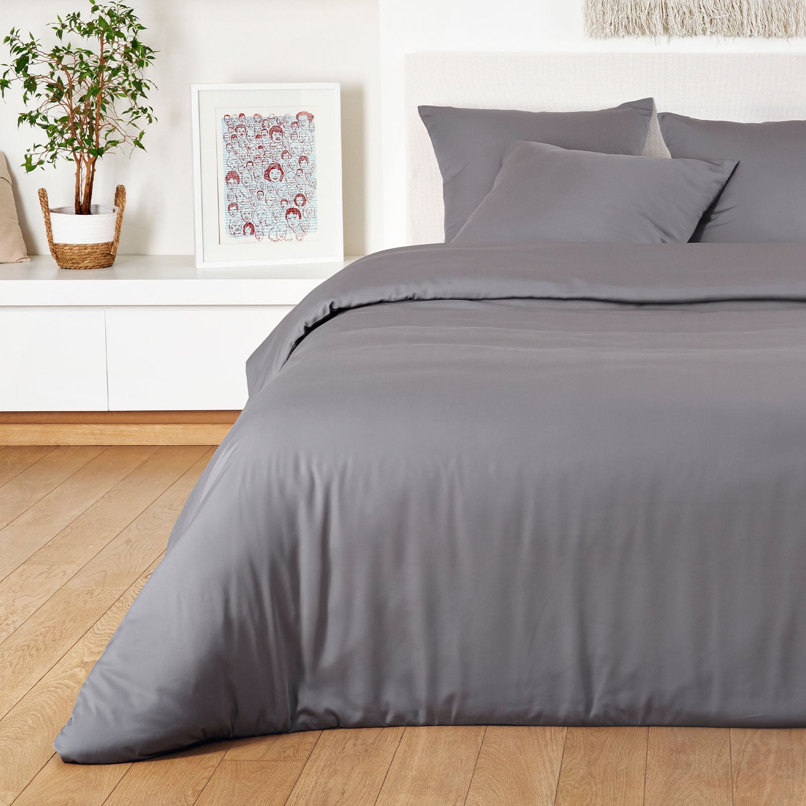 Gray bedding set on a bed in a bedroom with a plant and artwork on the wall.