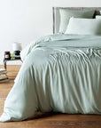 Bed with light green bedding in a room with a wooden floor and a nightstand.
