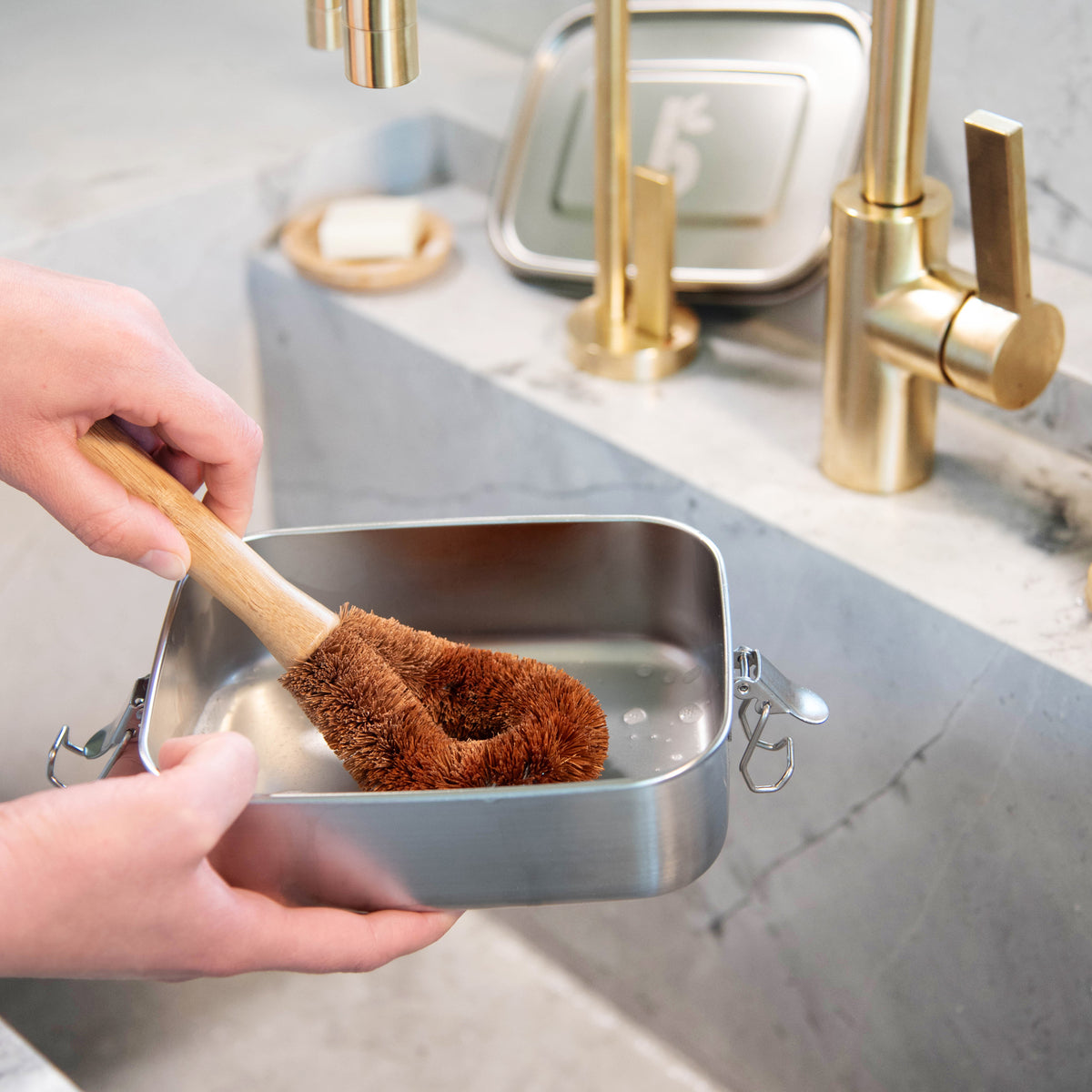Hand holding a wooden dish brush with brown bristles over a metal container on a kitchen counter.