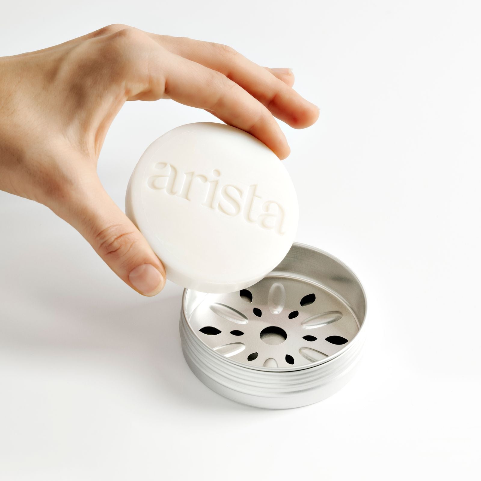 Hand holding a white 'arista' strengthening shampoo bar over a silver shampoo holder box on a light background