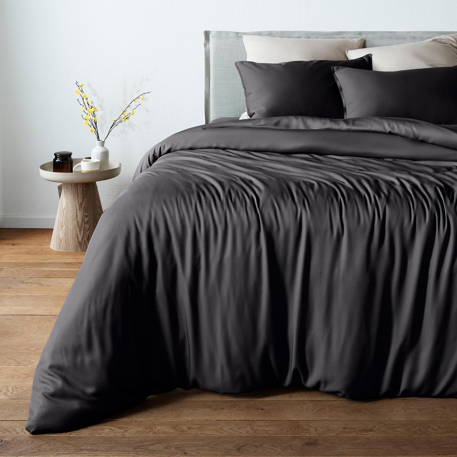 A bed dressed in black bambaw cozy bamboo duvet cover and matching pillow shams, styled in a bedroom with a light gray headboard, wood floor, and a small side table with a vase of yellow branches.