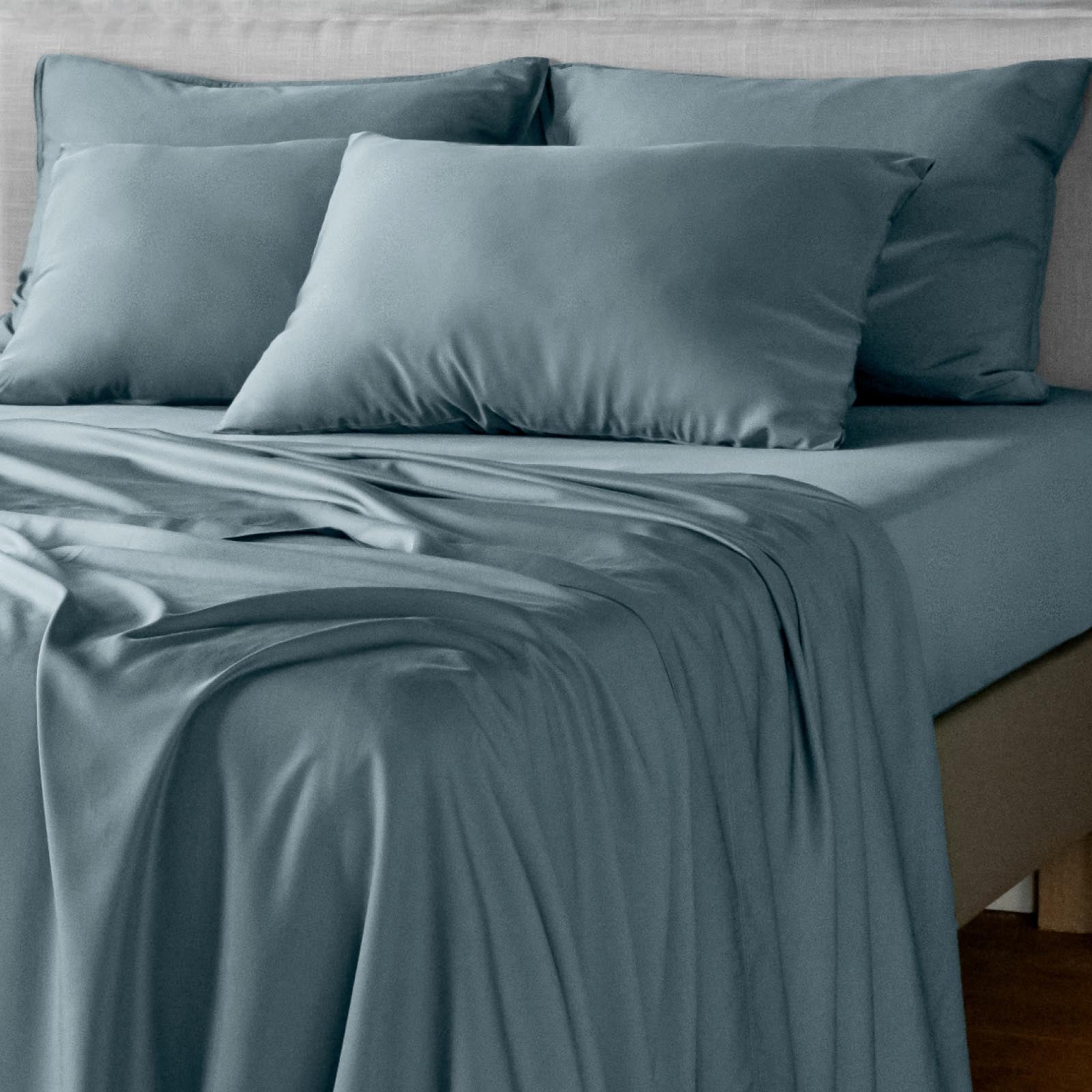 A neatly made bed with dusty blue bambaw cozy bamboo bedding, including a fitted sheet, pillowcases, and a matching duvet, styled with several pillows against a light neutral headboard.