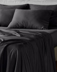 Black bamboo bed sheet and pillowcase set on a bed with rumpled fabric, shown in a dark studio setting.