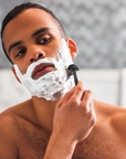A close-up lifestyle shot of a man shaving with bambaw metal safety razor white shaving foam on his face, holding a black razor against his cheek.