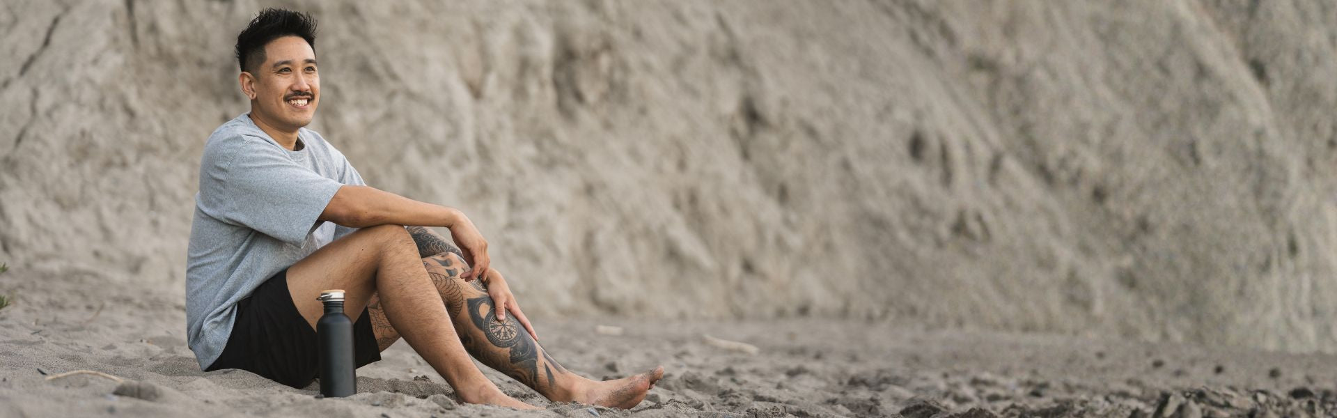 Man sitting on a sandy beach with a black bambaw stainless steel reusable water bottle